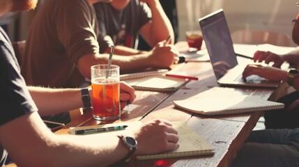 A group of people working together at a table with laptops and notebooks.  Sunlight streams in, illuminating the scene.  A glass of iced tea sits on the table.