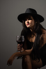 Beautiful woman in black clothes and hat leaning against the wall holding a glass of wine posing for the camera.