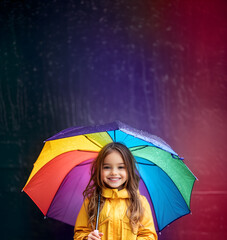 small 7 - 8 years old girl isolated in yellow raincoat holding colorful umbrella; smiled kid under the rainy weather; autumn/fall background; copy space