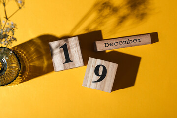 Wooden calendar with the date december 19 and flower in vase. Yellow background. Top view