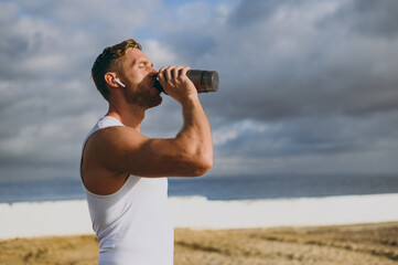 Young cool strong sporty athletic toned fit sportsman man wear sports clothes earphones drink protein shake warm up train at sunrise sun dawn over sea beach outdoor on seaside in summer day morning