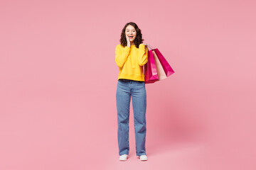 Full body young surprised woman wear yellow sweatshirt casual clothes hold in hand paper package bags after shopping isolated on plain light pastel pink background. Black Friday sale buy day concept.