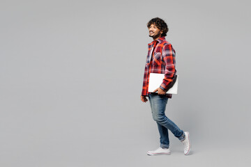 Side view young happy smiling Indian man he wearing red shirt casual clothes hold use closed laptop pc computer look aside isolated on plain grey color background studio portrait. Lifestyle concept.