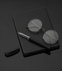 Black leather notebook with eyeglasses and pen on a black background