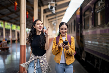 Two women are walking on a train platform, one of them is holding a cell phone