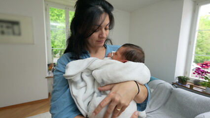 Mother cradling newborn baby, wrapped in white blanket, in modern living room, gazing lovingly at...
