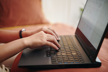 Hands typing on laptop keyboard while sitting comfortably on couch and working on calendar layout in a relaxed home environment showing engagement and productivity