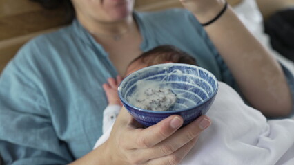 Mother holding a bowl of yogurt while newborn baby sleeps on her chest, multitasking and enjoying snack time, cozy indoor setting, intimate and nurturing family moment