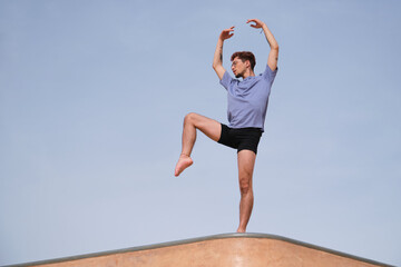 Young male ballet dancer performing choreography posing on building rooftop with blue sky