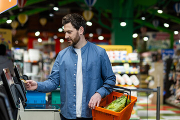 Man at self-checkout machine in grocery store holding orange basket with vegetables. Concept of modern shopping, technology use, convenience, and customer experience. Shopping with card payment. © Liubomir
