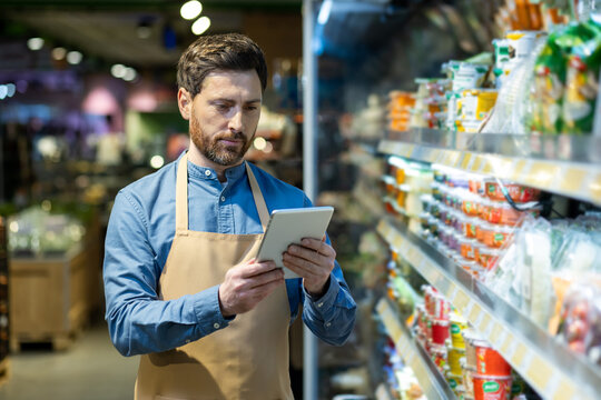 Grocery store employee with apron using tablet for inventory management in food aisle. Worker checking stock and organizing products in supermarket. Efficient retail operations with technology