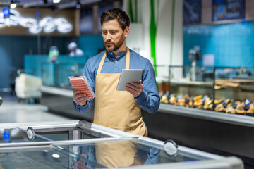 Supermarket employee wearing apron using tablet checking stock in grocery store. Man holding tablet...