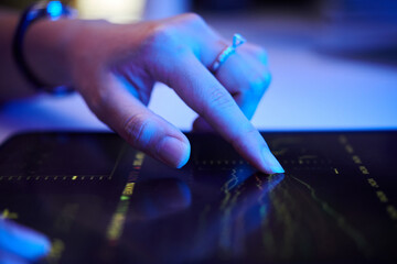 Close-up image showing hand with ring interacting with tablet screen displaying various financial data and charts under artificial lighting