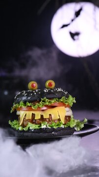 Monster burger. Black bun, beef cutlet, lettuce, onion, tomato and cheese in the shape of teeth, Against the backdrop of the moon and a steaming cauldron. Slow, vertical motion, circular rotation