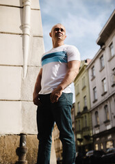 A man in a white shirt and blue jeans stands in front of a building. He is looking up at the sky