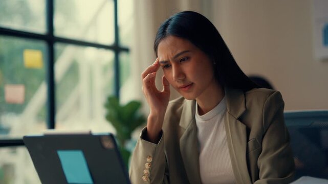 Businesswoman is overwhelmed with stress and a headache while working on a laptop in a modern office