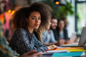 Fototapeta premium A focused woman with curly hair sits at a table among colleagues, engaged in a discussion or meeting with colorful papers and laptops in the background