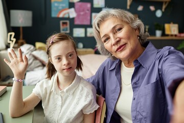 Portrait of senior woman and young girl with Down syndrome smiling and waving at camera in cozy home setting, surrounded by colorful decor without window