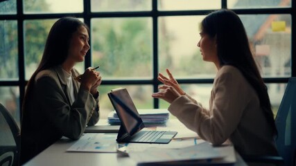 Two businesswomen discussing work at a desk in a modern office. Using a laptop and documents, they engage in a constructive conversation