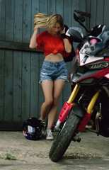 Plakat sexy beautiful blonde woman walking to her motorcycle, wooden wall in the background, looking thoughtfully at the ground