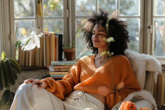 a happy woman sitting in her home office, relaxing with her eyes closed and smiling while doing yoga stretching on a chair near the window, wearing an orange sweater and white pants. Books are nearby - Powered by Adobe