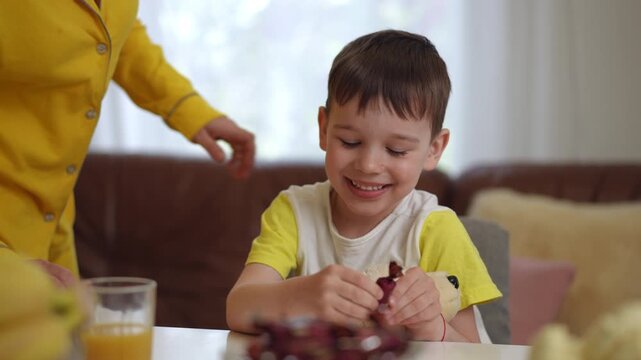 A smiling little boy, with a teddy bear, unwraps a candy in his hands, while sitting on a chair at a table in the living room. The mom walks up to the little boy and he laughingly, hides the candy