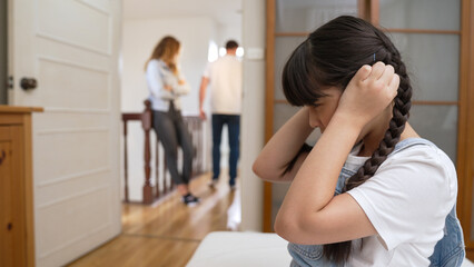 Stressed and unhappy young girl huddle in corner, cover her ears blocking sound of her parent arguing in background. Domestic violence at home and traumatic childhood develop to depression. Synchronos