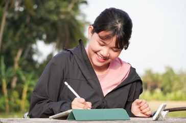 Happy young asian woman is working remotely at outdoor area or park using a tablet with smiling expression. Natural light with trees and nature background. Concept for freelancer.