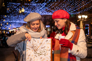 Girlfriends in coats and French berets with shopping bags enjoy cheerful Christmas shopping and a hot beverage at a festively decorated town square.