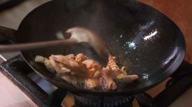 Adding soy sauce to the process of frying chicken fillet with onions with boiling oil, stirring with a ladle in a concave cast-iron frying pan on a gas stove, close-up.