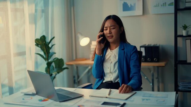 Businesswoman shows frustration and stress during a phone call in her office, surrounded by paperwork and a laptop