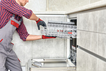Technician in uniform installs dishwasher into kitchen furniture. Repairman wear worker suit...