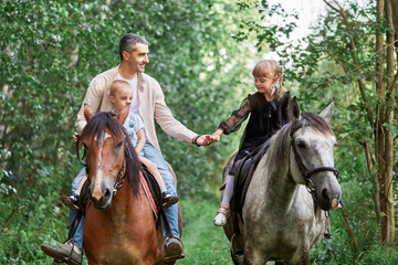 Happy dad rides horses with children in the forest in summer. A loving father spends time with his children, teaches riding, holds his daughter's hand.