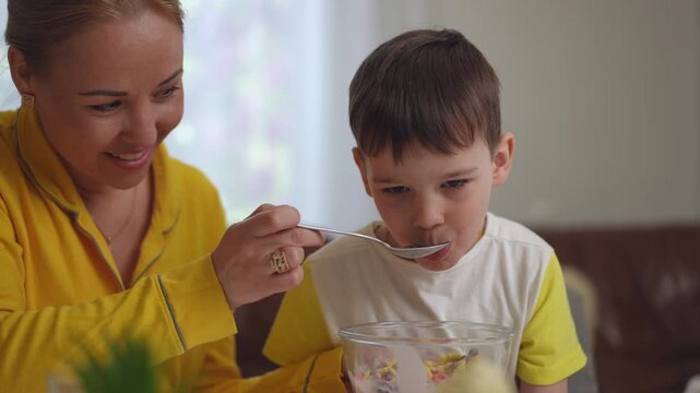 Close-up. A happy mother feeds her little son breakfast cereal and yogurt with a tablespoon, while sitting with him at the dining table in the living room of the house. The child is eating and