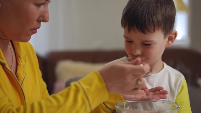 Close-up. A mother is trying to feed her small son breakfast cereal and yogurt with a tablespoon, while sitting with him at the dining table in the living room of the house. The child refuses to eat