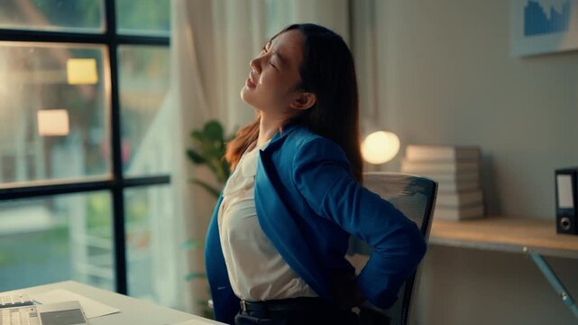 Businesswoman takes a break to stretch and relieve back pain while working on a laptop at her office desk. Bright workspace with a large window and office supplies