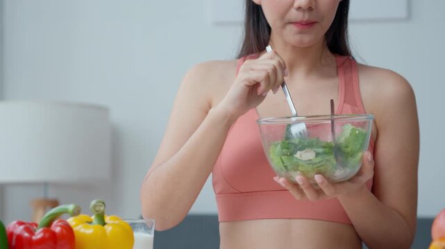 Young woman in sports attire prepares and eats fresh salad in a cozy kitchen surrounded by colorful vegetables. Ideal for health and nutrition themes