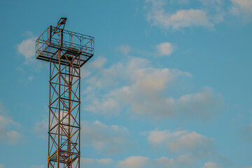 A tall metal tower with an observation platform stands against a clear blue sky dotted with soft clouds. The structure, made of metal beams, contrasts sharply with the tranquil sky