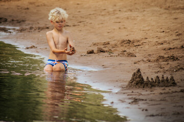 A young boy sits at the edge of a sandy shore, focused on building a sandcastle by the water. Wearing colorful swim trunks, he engages in creative play in the warm, peaceful setting. The calm water