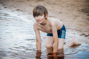 A young boy kneels in shallow water near the sandy shore, smiling softly as he engages in playful exploration. He wears blue swim trunks with colorful stripes, capturing the essence of a carefree © Emvats