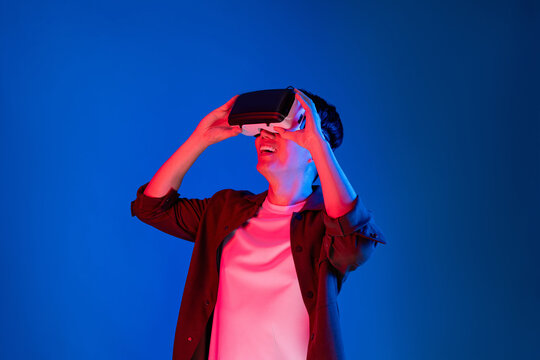 Asian man looking through VR enjoy watching fantastic meta on interesting object technology blue neon lighting background on virtual reality metaverse new world in 3D hologram innovation. Contrivance.