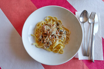 Overhead view of a white deep plate with tagliatelle carbonara pasta, with bacon, mushroom, onion, cream and basil. On a colorful tablecloth next to a stainless steel fork, knife and spoon and a paper