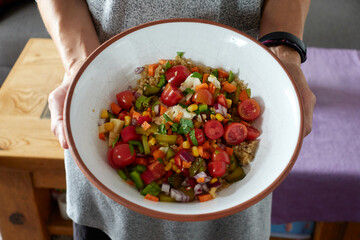 Woman hands showing a rustic ceramic bowl with quinoa salad with cherry tomatoes, pickles, red and green peppers, cheese, apple, corn, red onion, carrot and chopped cilantro