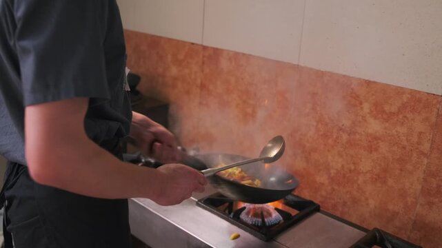 Young chef shaking wok concave pan throws up french fries and meat with vegetables fried on high heat with rising steam in restaurant kitchen.