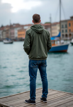 Backshot of a man standing on a dock in front of a body of water with a boat in the background and buildings on the other side of the water. Generative AI.