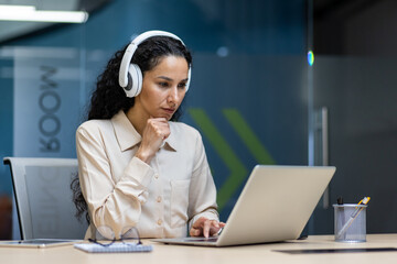 Focused woman wearing headphones working on laptop in modern office. Professional concentrating on task, using computer and technology. Concept of remote work, business, productivity