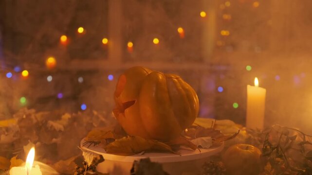 Halloween holiday background with terribly scary carved pumpkin blinks its eyes and mouth with a luminous lantern from the inside, rotating on a platform among garlands and candles in the smoke.