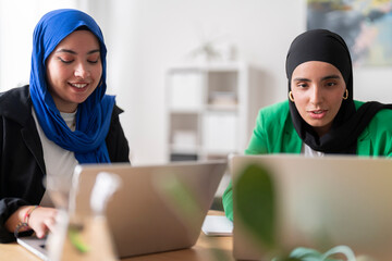 Two women in hijabs discuss work while sitting at a desk with laptops in front of them. The bright and modern office setting enhances their focused collaboration.