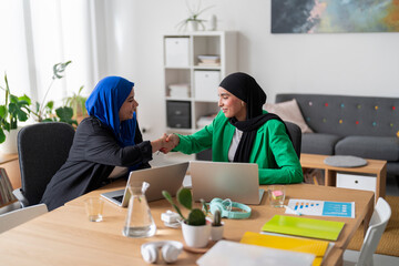 Two women in hijabs shake hands across a desk in a bright, modern office. Their friendly handshake signifies successful collaboration and agreement in a professional setting.