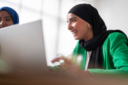 A woman in a black hijab and green blazer smiles as she works on her laptop in a bright office. Her positive demeanor adds to the professional yet friendly atmosphere of the workspace.